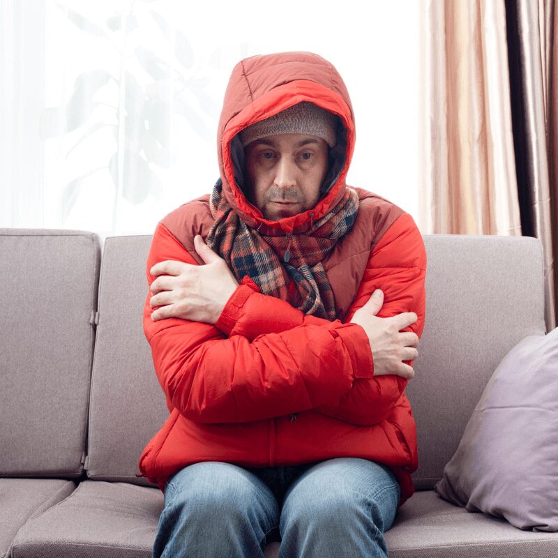 middle-aged white man sitting in winter coats on his couch because he’s cold.